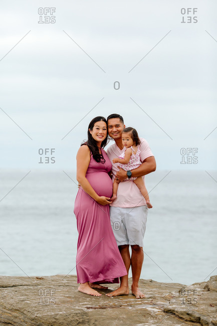 Beaming Asian dad, child and very pregnant wife on rocks by ocean