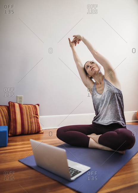 woman sits on yoga mat in front of laptop computer and stretches