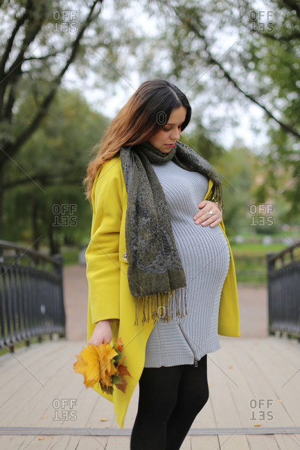 A pregnant woman stands in the park and looks at her belly.