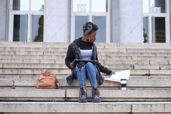 University female african student wearing protective face mask studying sitting on stairs outside on campus. New normal in college.