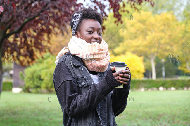 Beautiful female african woman wearing a scarf an holding a cup of coffee in a park. Fall, winter and cold concepts.