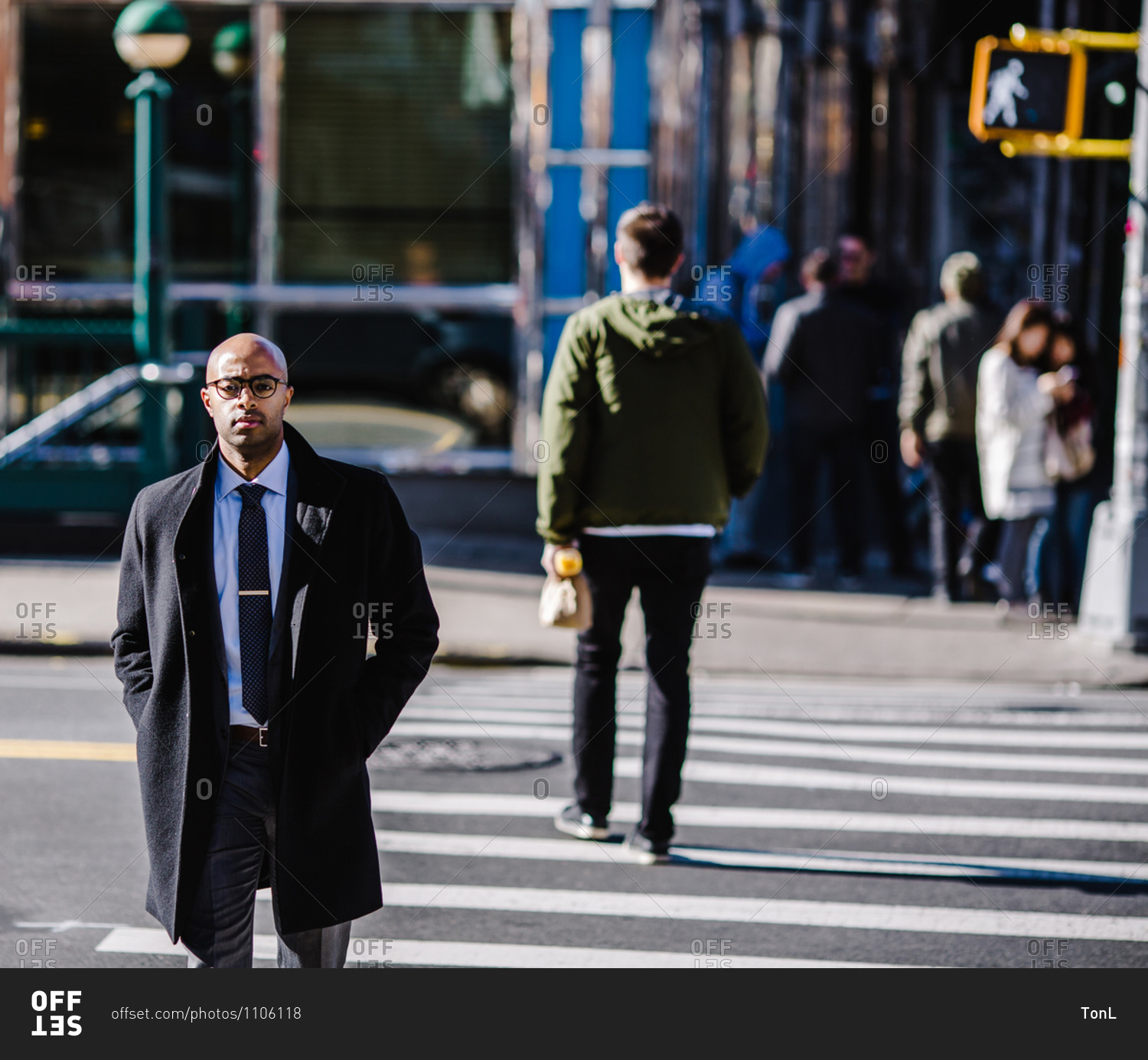 Medium shot of a bald black man in a heavy coat walking across a pedestrian crossing outside his office building