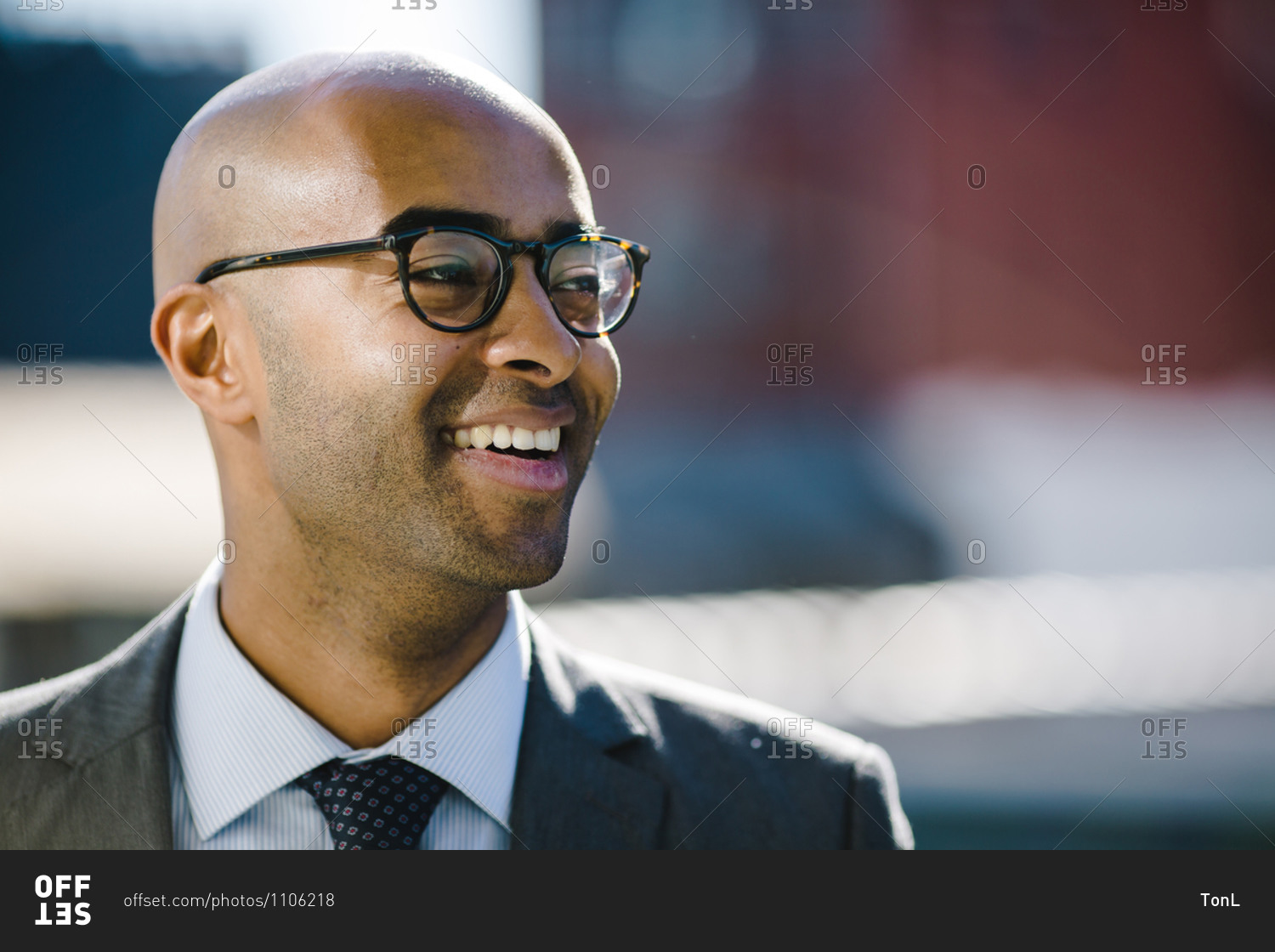 Portrait of a bald black man in a suit and glasses looking off into the distance and smiling