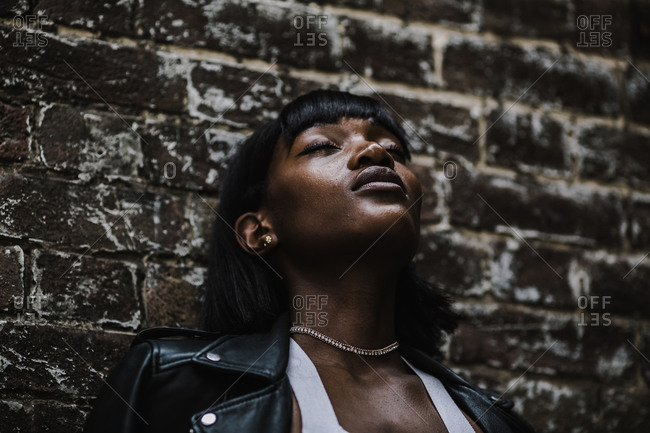 Closeup portrait of a black woman in a leather jacket leaning against a brick wall with her eyes closed