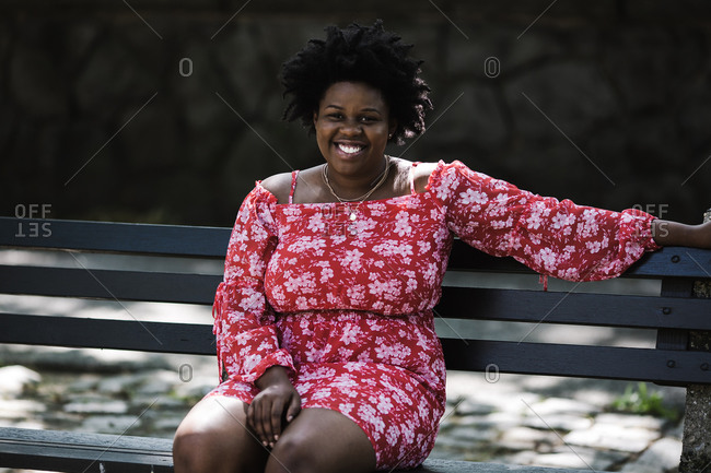 Black lady in a red floral dress smiling and sitting on a bench outdoors