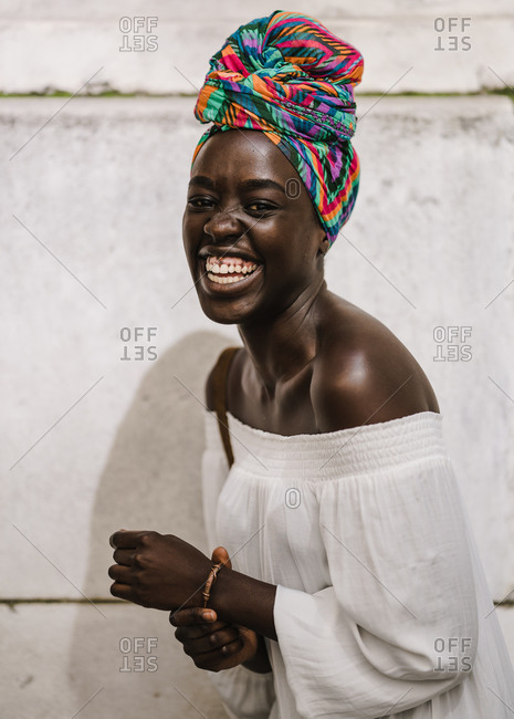 A portrait shot of dark skin black woman in a white off-shoulder blouse laughing while standing against a wall