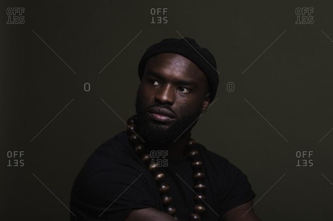 A close up shot of a serious African American man wearing a black beanie cap posing with a big beads necklace against a green background