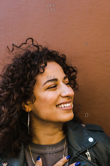 Vertical head and shoulder portrait of a curly hair latina woman in a black leather jacket in front of a brown wall smiles looking sideways