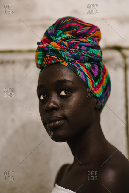 A close up shot of a dark skin black woman wearing a white off-shoulder blouse and a colorful headwrap against a wall