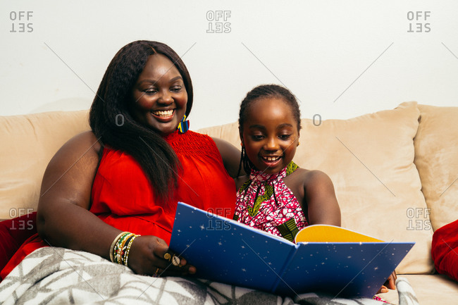 Black mother sitting on the couch and reading a book with her daughter