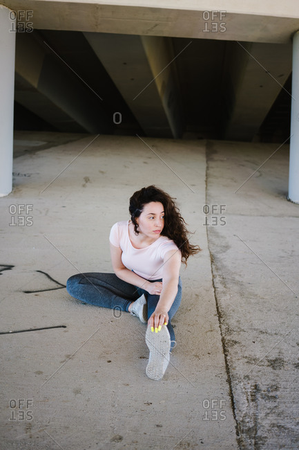 Young woman sitting on a slope under the bridge and doing warm up stretches