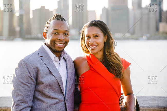 Waist up shot of a young black couple by a waterfront railing smiles at the camera
