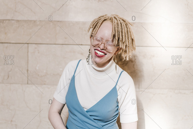 Horizontal waist up portrait of a joyous albino woman with eyes closed in front of a wall