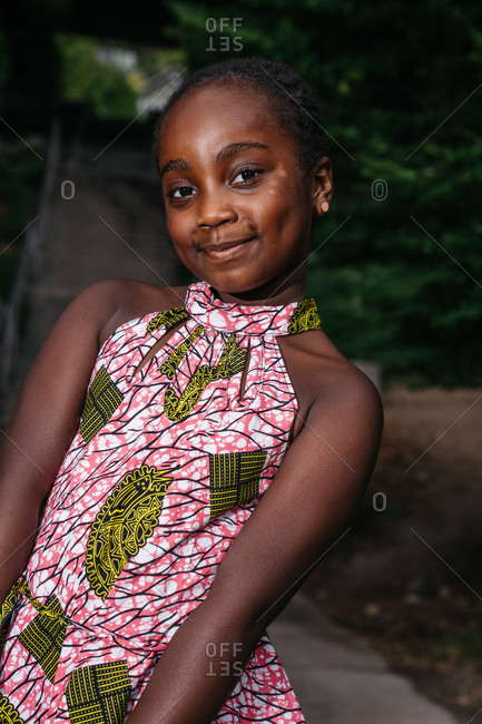 Vertical shot of a young Black girl wearing a floral dress and posing in front of a stairway in a park outdoors