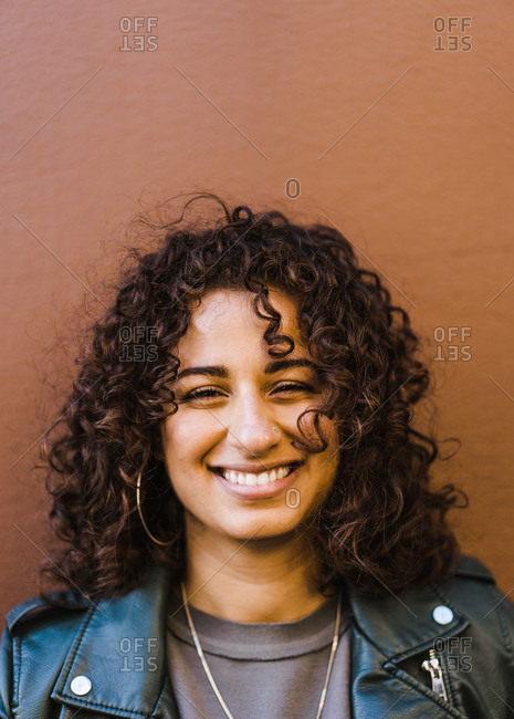 Vertical head and shoulder portrait of a curly hair latina woman wearing a black leather jacket in front of a brown wall smiles at the camera