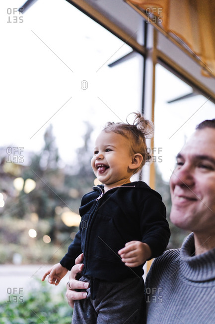 Vertical portrait of a father holding up his laughing daughter by the shoulder indoors