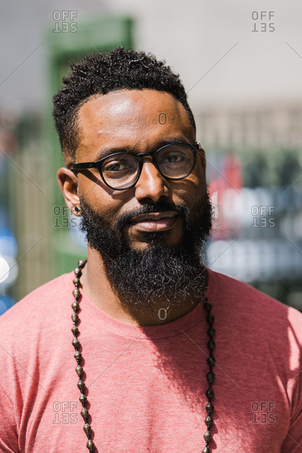 A close up shot of a black man with facial hair wearing glasses, red t-shirt, and necklace