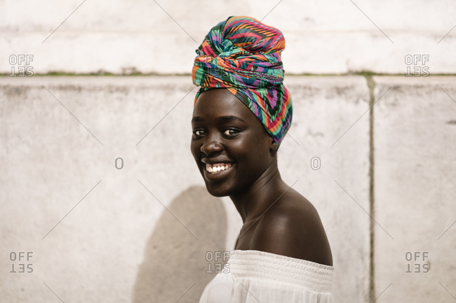 A close up shot of smiling dark skin black woman in a white off-shoulder blouse standing against a wall