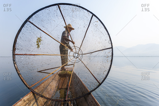 A basket fisherman on Inle lake scans the still and shallow water for signs of life and prepares to plunge his cone shaped net