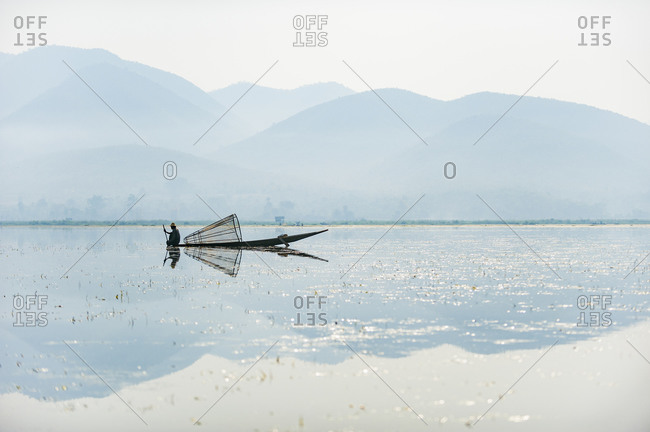 A basket fisherman on Inle lake scans the still and shallow water for signs of life and prepares to plunge his cone shaped net