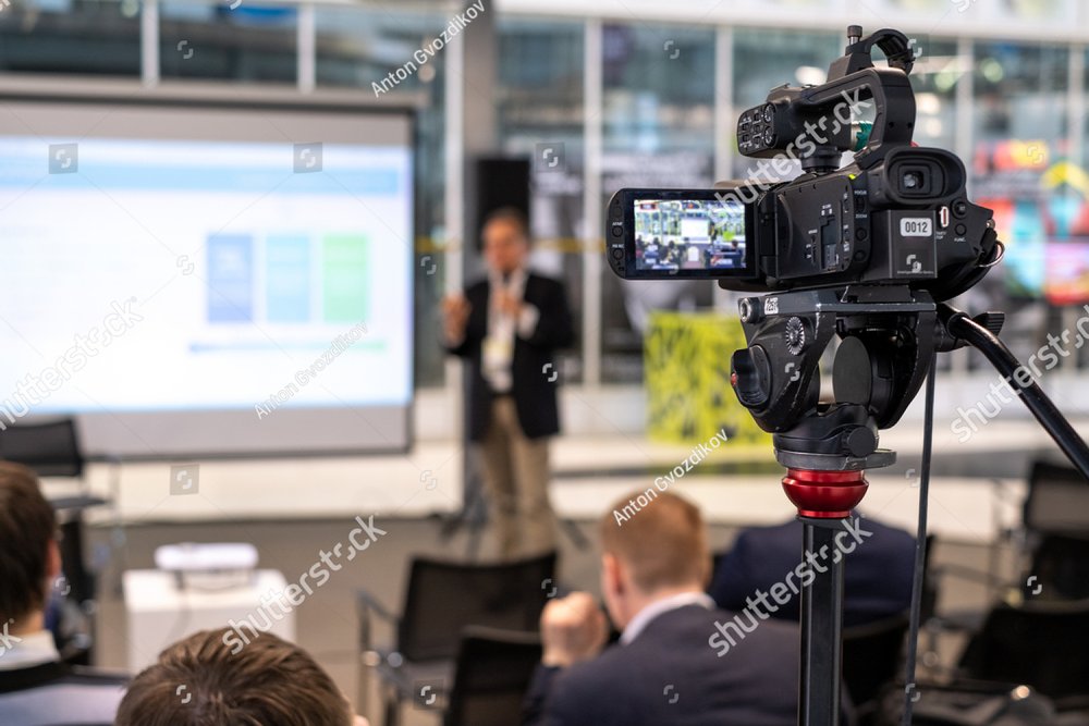 Video camera on tripod recording presentation of male coach standing on stage with display in front of audience