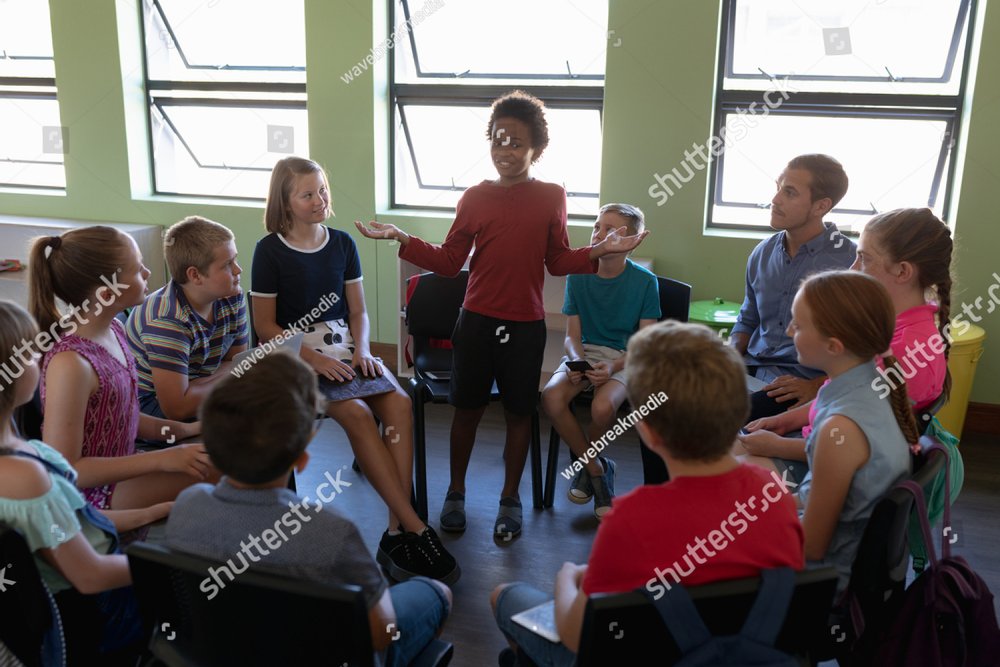 Front view of a diverse group of elementary school kids sitting on chairs in a circle and interacting during a lesson, one African American girl standing and talking while her classmates and male