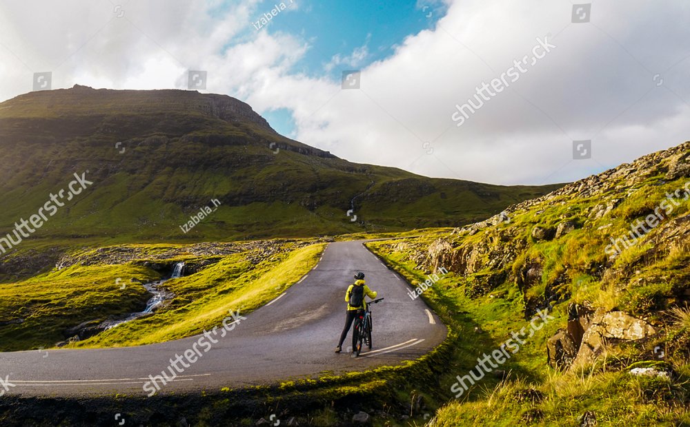 Female cyclist standing next to the bicycle on a mountain road. 
Undefined young bicyclist girl with a backpack, helmet and yellow coat.  Faroe Islands mountain biking in a beautiful natural scenery