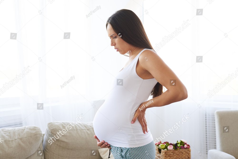 Side view of pregnant female suffering lower back pain, standing in living room 