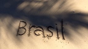 Brasil message handwritten on a smooth sand beach with palm tree shadows waving in the tropical wind