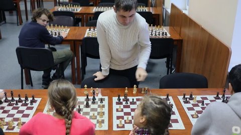 Five people play chess against one man in room with many chessboards on tables