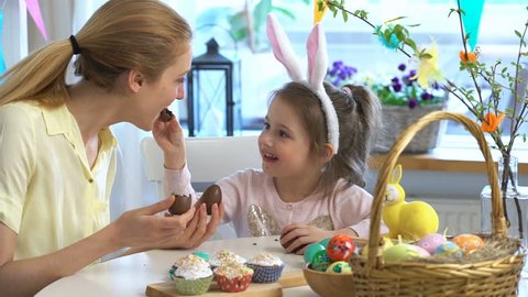 Mother And Daughter eating Chocolate Easter Eggs sitting at the holiday table with a basket and a yellow rabbit. Slow Motion