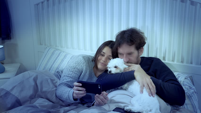 Couple in pajamas in bed taking selfie with puppy dog at night