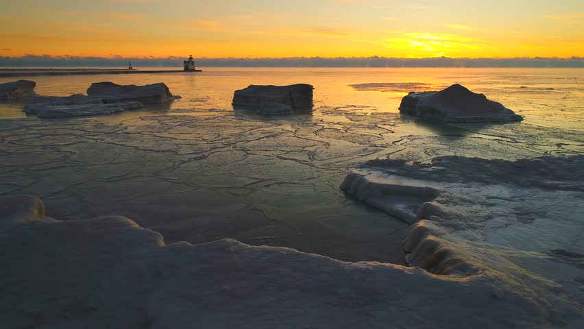 Winter dawn sky over frozen Lake Michigan shoreline with icebergs, sea smoke, lighthouse in distance, mallards suddenly fly by!  Moving aerial shot from drone.