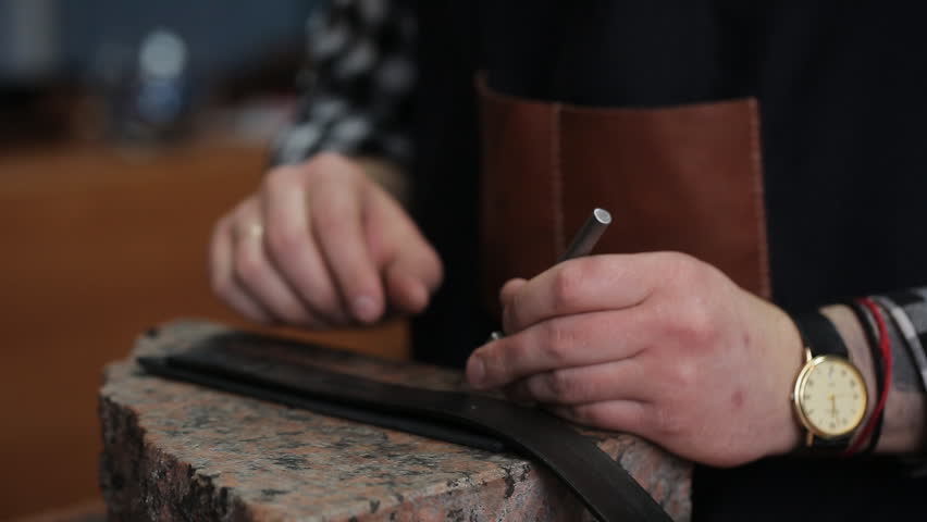 Working process of the leather belt in the leather workshop. Man holding crafting tool and working. He is placing billet to sturdy stand, fixing round knife on a mark and hitting with a small hammer Royalty-Free Stock Footage #1006623397