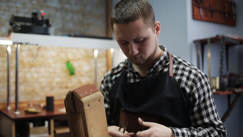 Working process of the leather belt in the leather workshop. Man holding crafting tool and working. He is placing billet to sturdy stand, fixing round knife on a mark and hitting with a small hammer Royalty-Free Stock Footage #1006623412