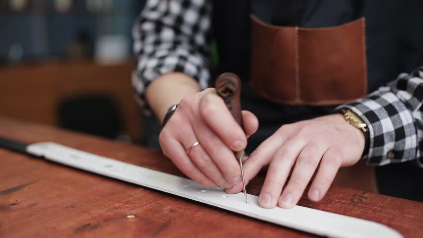 Working process of the leather belt in the leather workshop. Man holding crafting tool and working. He is placing billet to sturdy stand, fixing round knife on a mark and hitting with a small hammer Royalty-Free Stock Footage #1006623418