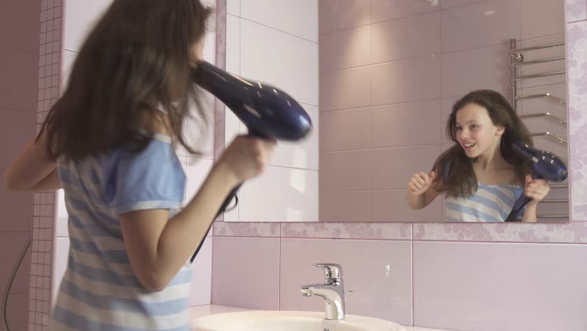 Beautiful happy girl teenager dries hair with a hair dryer and sings and dances in front of a mirror in the bathroom stock footage video