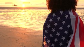 4K video clip of mixed race African American girl teenager female young woman wrapped in an American US Stars and Stripes flag, watching people playing on a beach at sunset or sunrise
