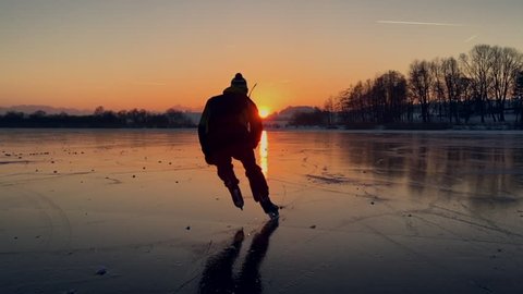 Panoramic slow-motion view of a young hockey player on a frozen lake skating into the sunset with amazing reflections during beautiful golden evening light in winter