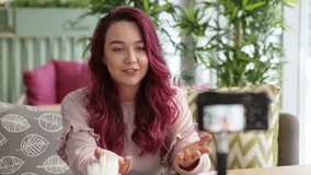 Young attractive female blogger with pink fashion hair blogging in cafe. Happy smiling woman with camera recording video tells about life, lifestyle. Technology, videoblog, mass media concept.