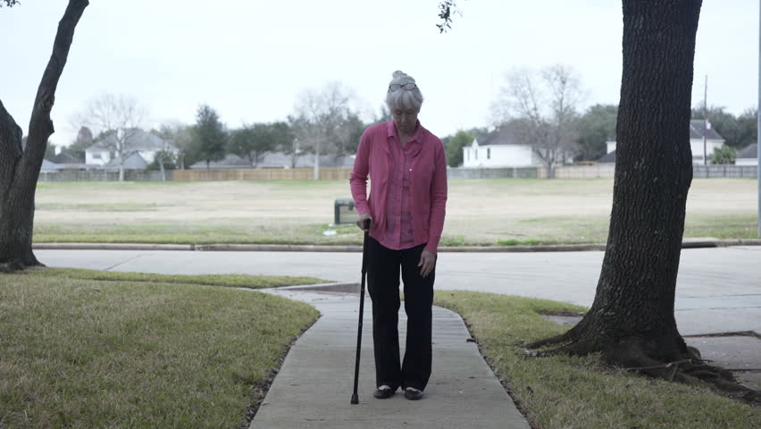 A Caucasian senior citizen woman with a cane walking slowly and carefully on a neighborhood sidewalk.