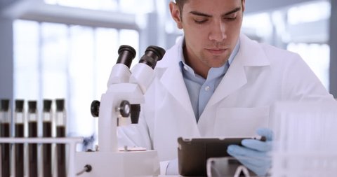 Young Hispanic male scientist in laboratory using tablet. Serious medical researcher looking through microscope. 4k
