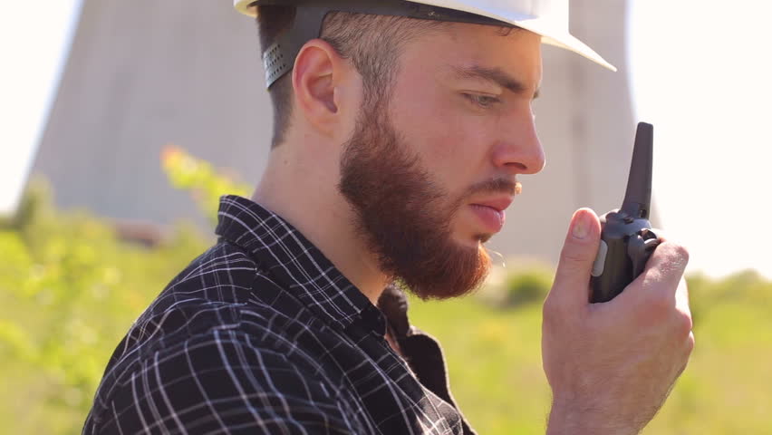 Close-up of a bearded man Builder in a protective white helmet talking on the radio in the background of large industrial pipes.