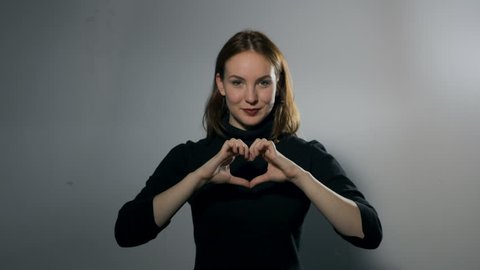 A young woman standing behind a white wall making a heart hand gesture that turns into a broken heart hand gesture and smile to a frown as a consequence. Medium close up.