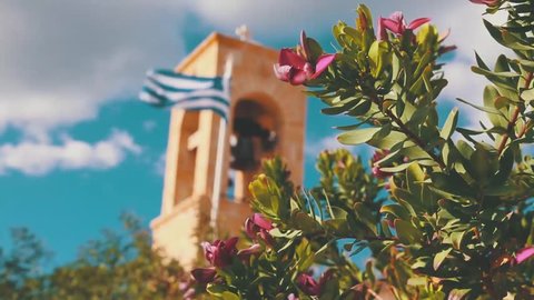Greek church bell and flag in the wind on a sunny day, blooming pink flowers on foreground