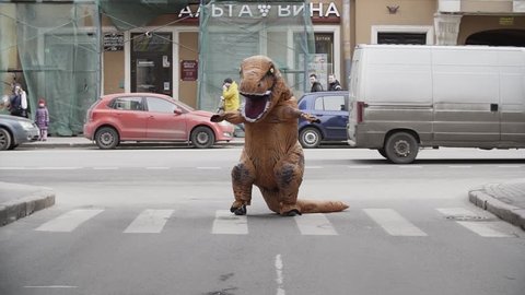 SAINT PETERSBURG, RUSSIA - APRIL 1, 2017: Man in dino mascot jumping and dancing at city street crosswalk with lots of cars. Sunny day. Slow motion