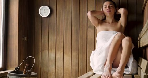 A beautiful woman wearing a white towel takes a sauna: The sauna is made of wood with a large window with a view of the snow. Concept of: relax, vacation, wellness center.