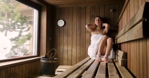 A beautiful woman wearing a white towel takes a sauna: The sauna is made of wood with a large window with a view of the snow. Concept of: relax, vacation, wellness center.