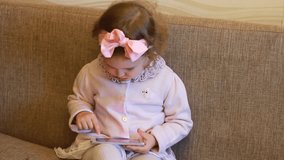 A little cute girl is sitting in sofa in a living room, looking cartoon and playing the game on a smartphone. Funny child looks on the mobile phone screen and plays downloaded application