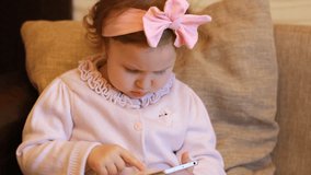 Funny child looks on the mobile phone screen and plays downloaded application. A little cute girl is sitting in sofa in a living room, looking cartoon and playing the game on a smartphone.
